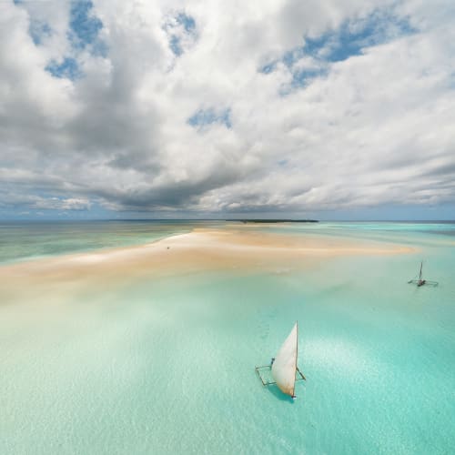 Ein Segelboot schwimmt ruhig auf klaren türkisfarbenen Gewässern in der Nähe einer Sandbank mit einer entfernten Insel und einem bewölkten Himmel.