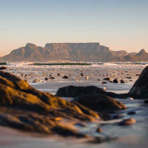 Blick auf den Tafelberg von einem Strand aus, mit Felsen und nassem Sand im Vordergrund während des Sonnenuntergangs.