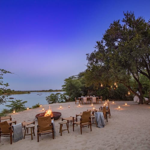 Ein gemütliches Outdoor-Ess-Setup mit Stühlen rund um ein Lagerfeuer am Sandufer eines Flusses bei Sonnenuntergang, im Hintergrund Bäume und Wasser.