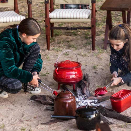 Zwei Mädchen kochen im Freien am Lagerfeuer, umgeben von Stühlen und Kochgeschirr.