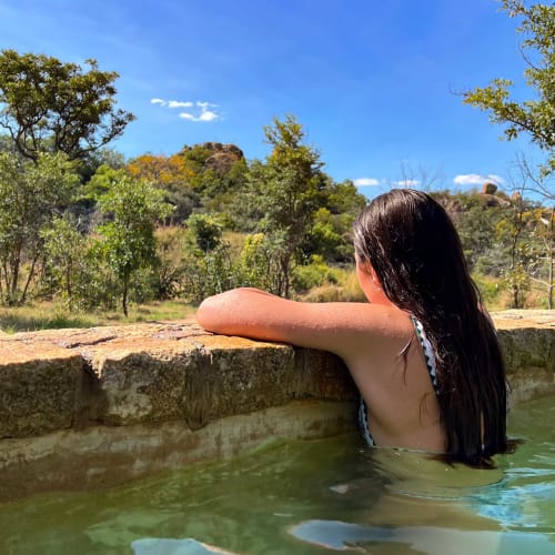 A woman with long dark hair relaxing in a natural-looking pool with stone edges, surrounded by green trees under a bright blue sky.