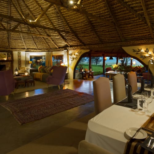 Interior of a cozy, rustic lounge and dining area with a thatched ceiling, large windows showing a green landscape, and elegant furniture and table settings.