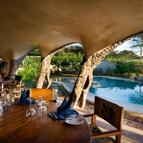 Indoor dining area with a view of a swimming pool and lush greenery outside, featuring wooden furniture and a curved ceiling. 