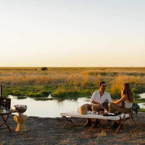 Ein Paar macht ein Picknick am Flussufer bei Sonnenuntergang in einer flachen Graslandschaft.