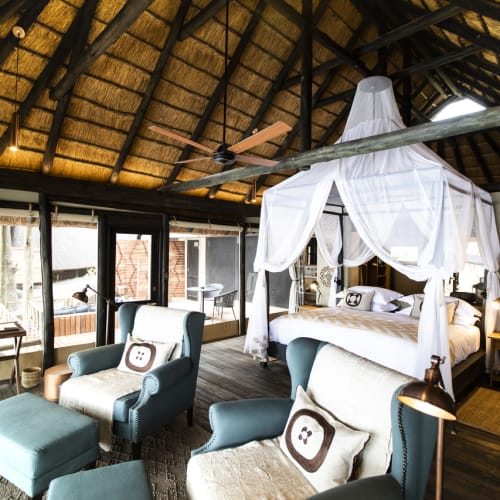 Luxurious bedroom with a canopy bed in a thatched-roof hut, surrounded by seating and large windows.