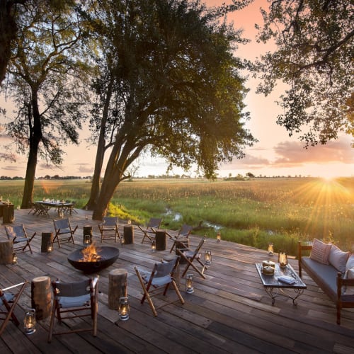 A cozy outdoor seating area on a wooden deck with a fire bowl, surrounded by trees and overlooking a sunset in a grassland.
