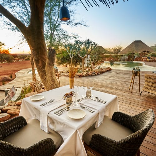 A luxurious outdoor dining setup on a wooden deck overlooking the Kalahari landscape at sunset, with a pool in the background.