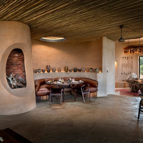 Elegant dining area with a curved leather banquette, wooden chairs, and decorative pottery on a ledge, illuminated by natural light from a window.