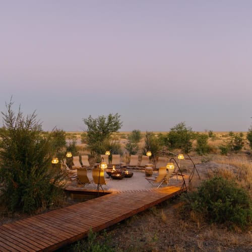 A cozy campfire setup in an outdoor natural setting with chairs and tables around a fire pit, surrounded by sparse bushes and trees in a flat landscape.