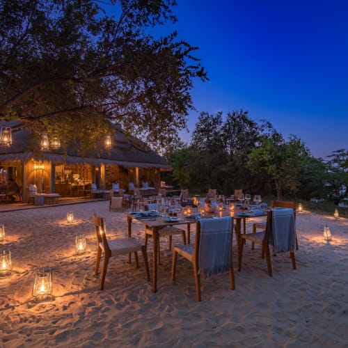 A romantic outdoor dinner setup on a sandy surface at dusk with candle-lit tables and hanging lanterns under trees.
