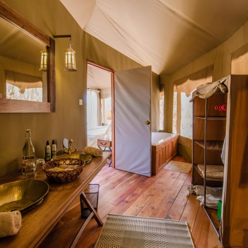 Innenraum eines luxuriösen Zeltes mit Holzmöbeln, einem Spiegel und einem Bett, sichtbar durch eine offene Tür.