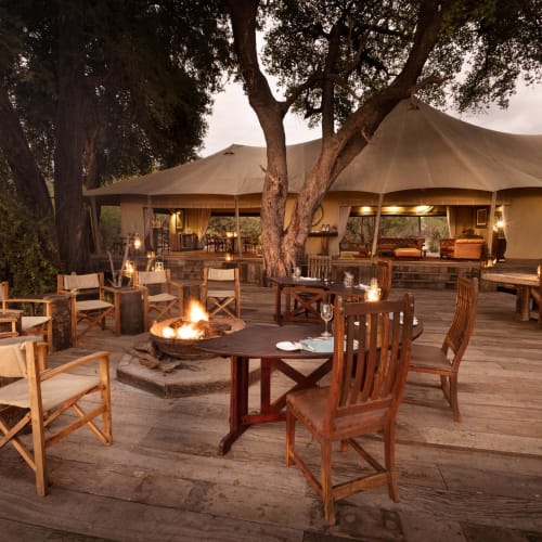 A cozy outdoor gathering area with wooden chairs around a fire pit under large trees and a tent structure in the background.