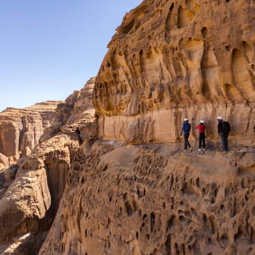 Eine Gruppe von Menschen wandert entlang eines schmalen Weges, der in bunte, geschichtete Felsformationen in Alula gehauen wurde. Die Landschaft zeigt raue Klippen und einen klaren blauen Himmel.
