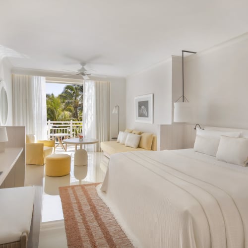 Eine helle Hotelsuite mit großem Bett, Sitzbereich und Balkon mit Blick auf tropische Bäume.
