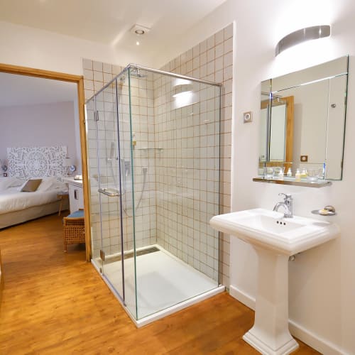 Badezimmer mit einer duschkabine aus Glas, einem weißen Standwaschbecken mit Spiegel darüber und Holzboden, neben einer offenen Tür, die zu einem Schlafzimmer mit Bett und dekorativem Kopfteil führt.