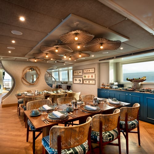 Innenraum eines Speisesaals auf einem Kreuzfahrtschiff mit einem eingedeckten Holztisch und moderner Dekoration, Blick auf den Ozean durch ein großes Fenster.