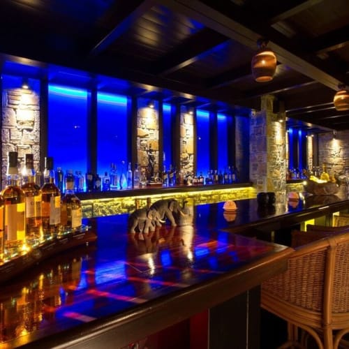 Eine elegante Bar mit Flaschen von Spirituosen auf dem Regal, beleuchtet mit blauem und gelbem Licht, mit Korbsesseln entlang des Counters in einem stilvollen, schwach beleuchteten Innenraum.