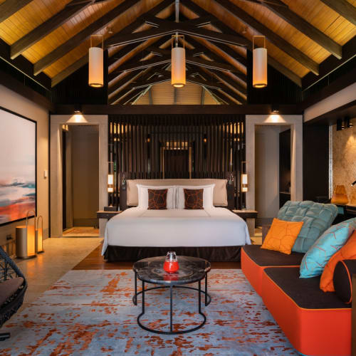 Luxuriöses Schlafzimmer in einer Strand-Pool-Villa mit gewölbter Holzdecke, großem weißen Bett mit Kissen, orange-schwarzem Sofa mit bunten Kissen, Hängesessel und moderner Beleuchtung.