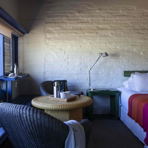 Cozy hotel room with a large bed, a round table, chairs, and a window showing trees outside, in a room with white brick walls.