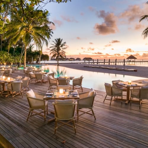 Freiluftrestaurant mit Tischen und Stühlen auf einer Holzplattform, mit Blick auf einen Strand mit Palmen und Sonnenuntergang im Hintergrund.