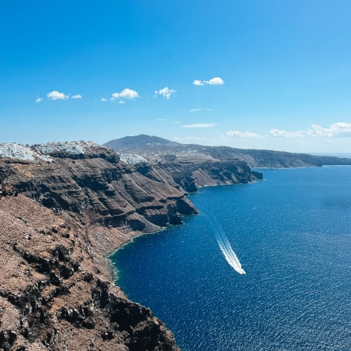 Deep blue waters of the Aegean Sea bordered by the rocky cliffs of Santorini's caldera with a small boat leaving a white trail.