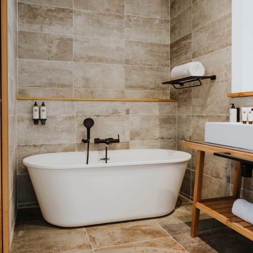 Modernes Badezimmer mit einer freistehenden weißen Badewanne vor großen beigen gefliesten Wänden, einem schwarzen Duschkopf und Wasserhahn, einem hölzernen Waschbeckenunterschrank mit weißem Waschbecken sowie ordentlich angeordneten Toilettenartikeln und Handtüchern.