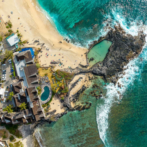Luftaufnahme eines Küstenstrands mit türkisfarbenem Meer, sandigem Strand mit Menschen, Felsformationen und einem Meerwasserbecken in der Nähe von Strandhäusern und Bäumen.