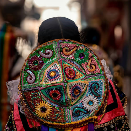 A person wearing a traditional, colorful embroidered hat with ribbons, seen from behind in a market setting.