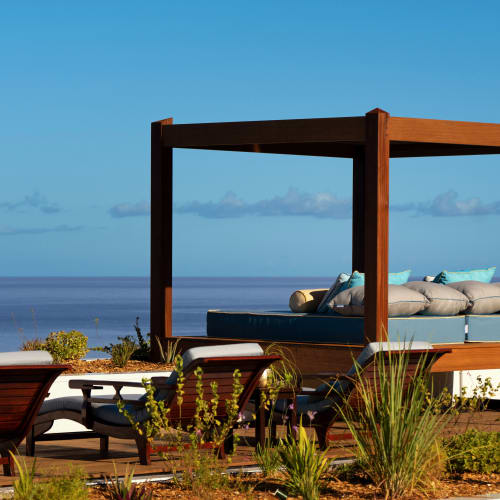Ein luxuriöses Außentagesbett mit Kissen unter einem Holzdach mit Blick auf einen ruhigen blauen Ozean und klaren Himmel, umgeben von Pflanzen und Terrassenmöbeln.