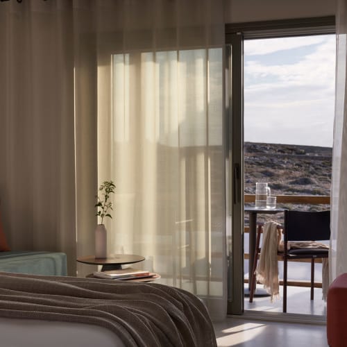 Sunlit hotel suite interior with bed, bedside table, plant vase, and seating area, opening to a balcony overlooking a rocky landscape under a cloudy sky.
