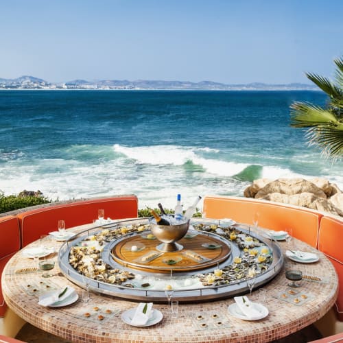 Ein luxuriöser runder Tisch im Freien mit Geschirr und Gläsern, blickt auf das Meer mit Wellen und einer Palme.