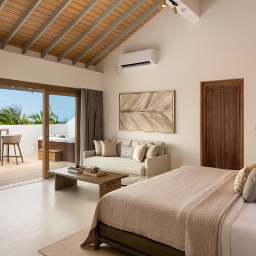 Modernes Hotelzimmer mit großem Bett in beigem Leinen, beigem Sofa, Holztisch und Schiebetüren, die auf einen Balkon mit Stühlen und üppigem Grün draußen öffnen.