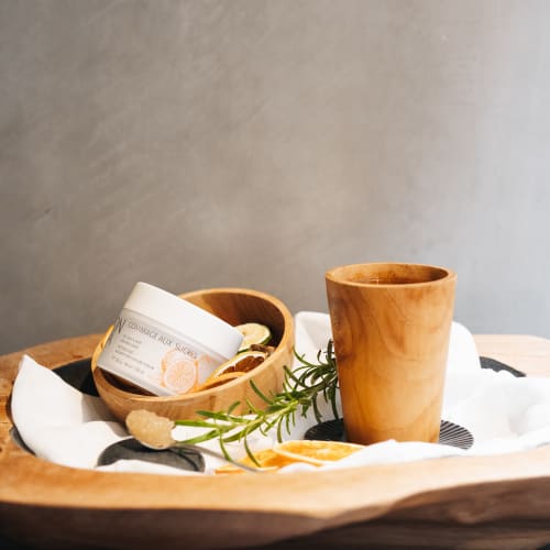 Ein Holzbecher und eine kleine Holzschale mit einem Hautpeeling-Behälter, auf einem weißen Tuch mit einem Rosmarinzweig und getrockneten Zitrusscheiben auf einem hölzernen Tablett.