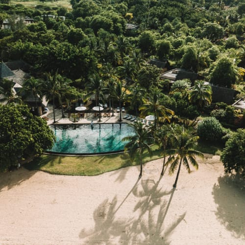 Luftaufnahme eines tropischen Resorts mit Strand, Palmen, die lange Schatten auf den Sand werfen, einem Schwimmbecken mit Sonnenschirmen und dichtem grünem Wald, der bis zu den Bergen im Hintergrund reicht.