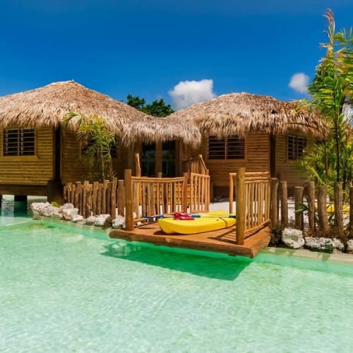 Zwei strohbedeckte Holzhäuser auf Stelzen über einem klaren, türkisfarbenen Wasserpool mit Holzsteg und gelben Kajaks, umgeben von grünen Palmen unter einem blauen Himmel.