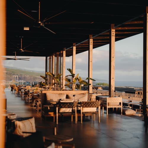 Überdachte Außenterrasse mit Holzstühlen, Tischen und gepolsterten Bänken mit Blick auf eine malerische Küstenlandschaft im warmen Abendlicht.