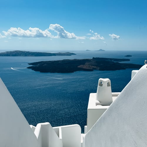 White stairway leading down towards a deep blue sea with volcanic islands under a bright blue sky with scattered clouds in Santorini, Greece.