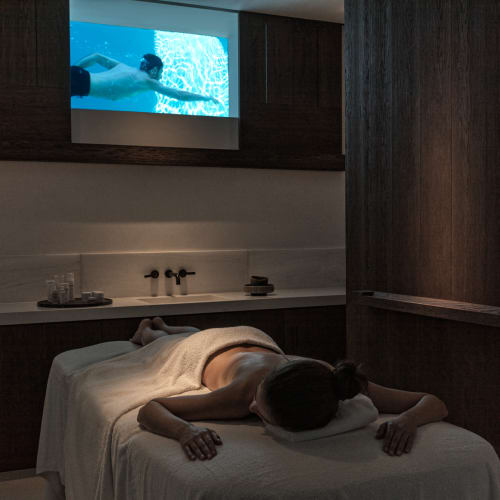 Person liegt mit dem Gesicht nach unten auf einer Massageliege in einem schwach beleuchteten Spa-Raum mit einem Bildschirm, der eine Unterwasserschwimmszene zeigt.