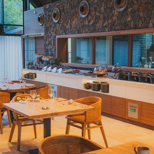 Gemütlicher Essbereich in einem Restaurant mit Holztischen und Stühlen, die für Mahlzeiten gedeckt sind, mit einem Buffet-Tresen an einer Wand mit Steinakzenten und großen Fenstern, die natürliches Licht hereinlassen.