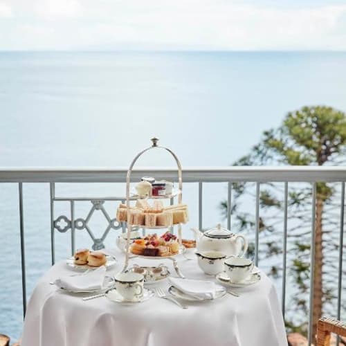 Elegante Nachmittagstee-Einrichtung mit einem Tisch voller Teeservices und Desserts mit Blick auf das Meer von einem Balkon.