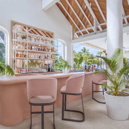 A stylish bar area with a curved pink counter, surrounded by pastel pink chairs and large potted plants, with big windows showing a tropical outdoor setting.