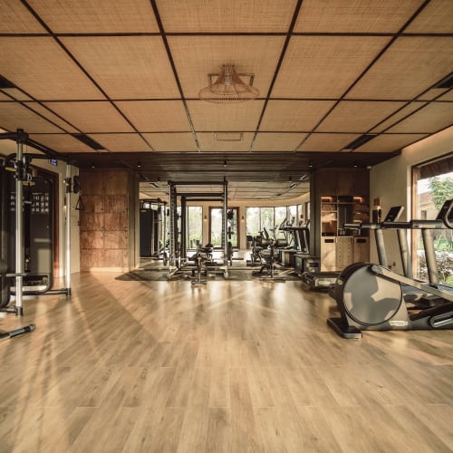 Large gym room with wooden flooring and ceiling, filled with various exercise machines including treadmills, rowing machines, and weight equipment, surrounded by windows revealing greenery outside.