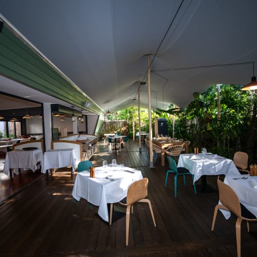 Außenbereich eines Restaurants mit Tischen mit weißen Tischdecken und modernen Stühlen unter einem großen Zeltdach umgeben von grünem Laub.