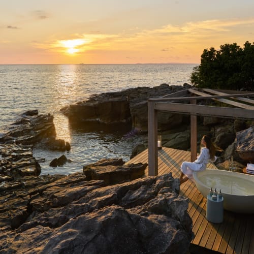 A woman relaxing in a bathtub on a wooden deck by the rocky coastline at sunset, with the ocean in the background.