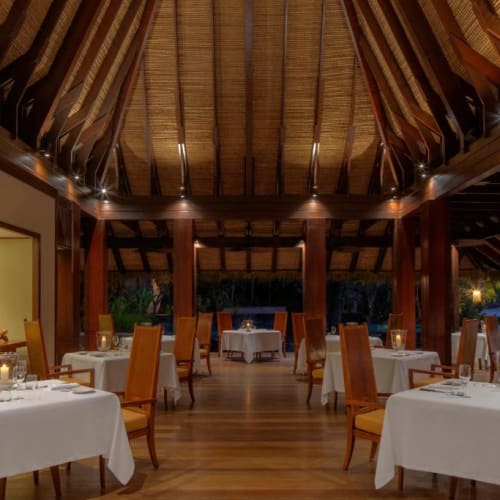 Elegantes Restaurant mit runden Tischen und orangefarbenen Stühlen unter einem hohen Holzdach bei Sonnenuntergang.