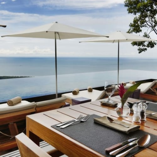 Eleganter Außenbereich mit Esstisch am Hotel mit Blick auf den Ozean, Sonnenschirmen und Bäume.