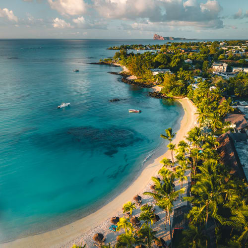 Aerial view of a curved sandy beach lined with palm trees and sunbeds, next to calm turquoise waters with a few boats, and a green coastal town stretching into the distance under a partly cloudy sky.