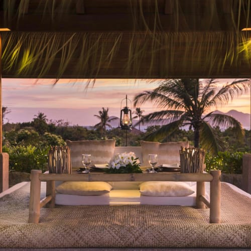 A romantic dinner setup on a raised bed with cushions, overlooking a tropical landscape during sunset.