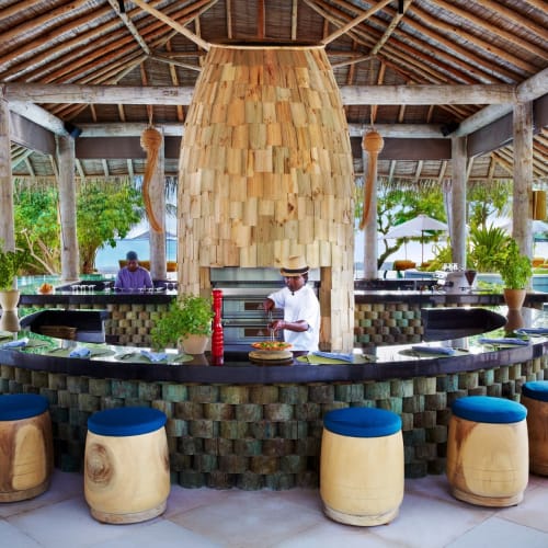 Ein Barkeeper steht hinter einer runden Bar mit bunten Hockern in einem Open-Air-Resort-Restaurant, umgeben von üppigem Grün und Palmen.