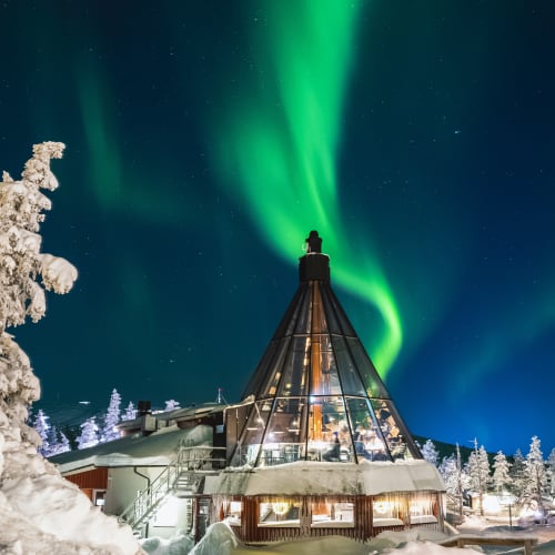 Crowne Iglu Restaurant with a triangular glass structure surrounded by snow-covered trees under a vibrant green Northern Lights sky at night.